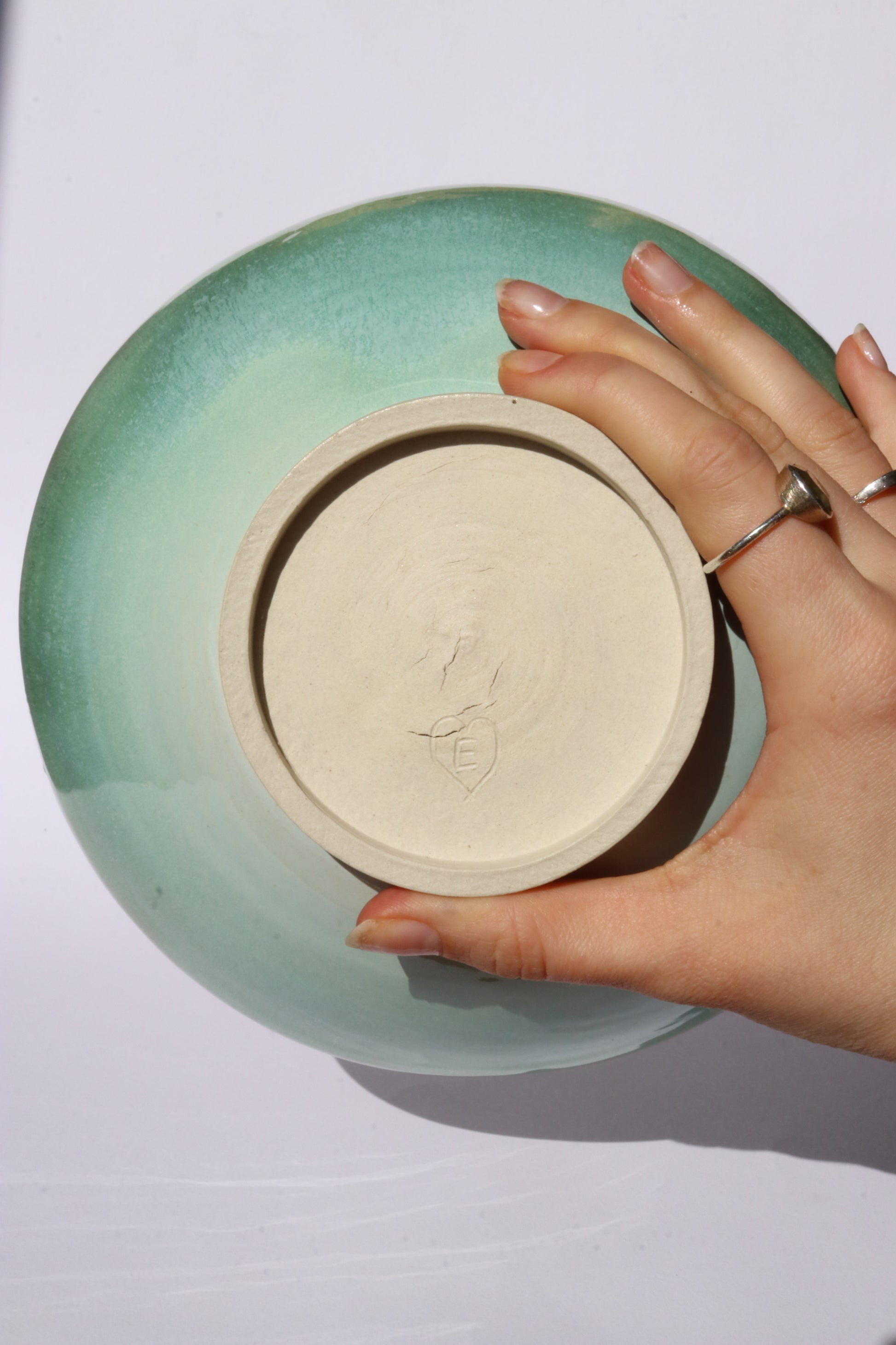 Hand holding a ceramic plate with a green rim against a white background, showing the bottom and the signature